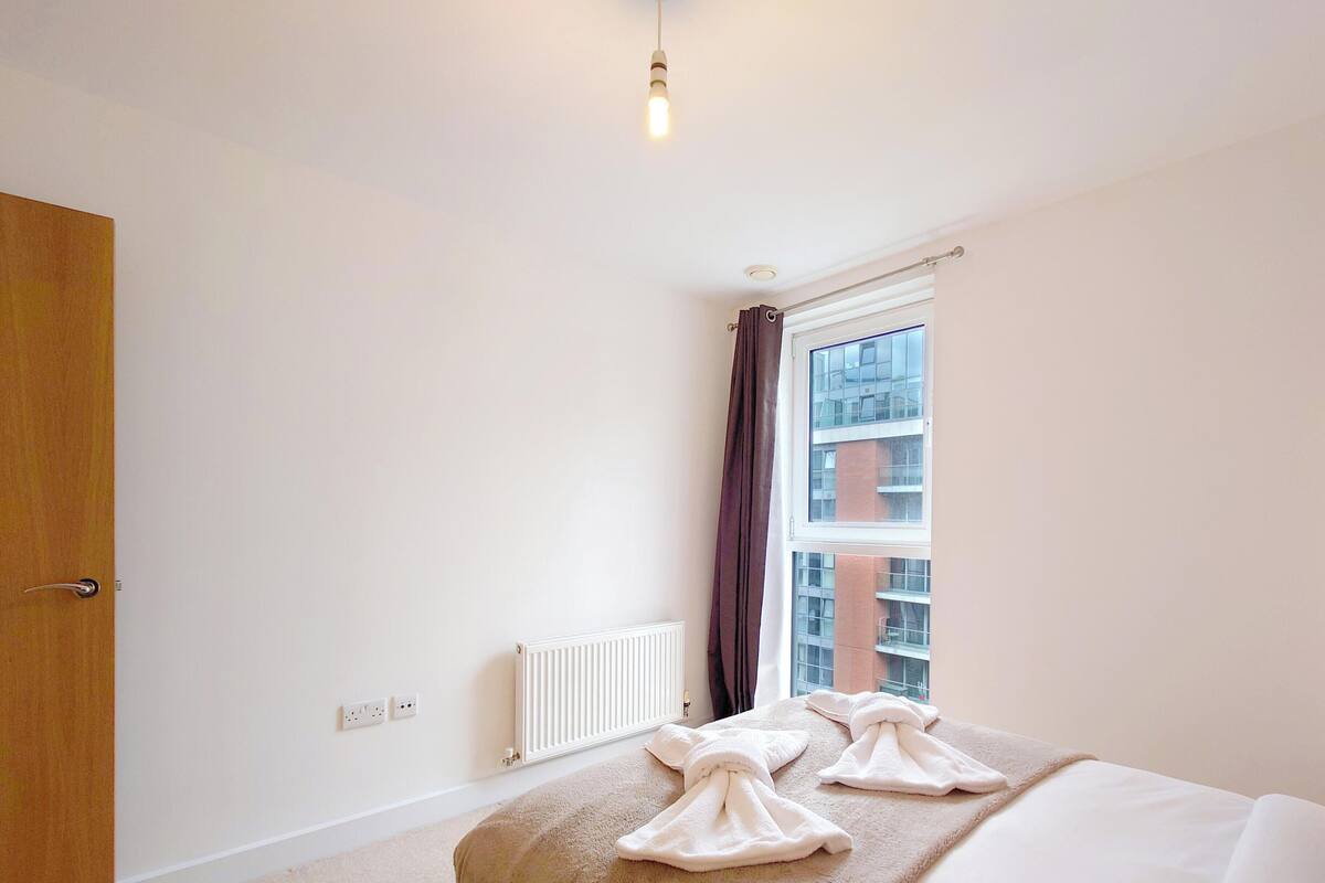 Un dormitorio con cama, ventana con cortinas, radiador y lámpara.