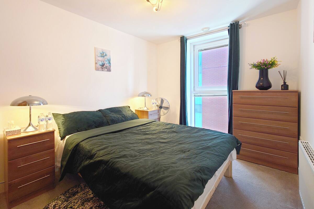 Un dormitorio con cama, mesitas de noche, cómoda, ventana con cortinas, ventilador de pared y una pequeña obra de arte.