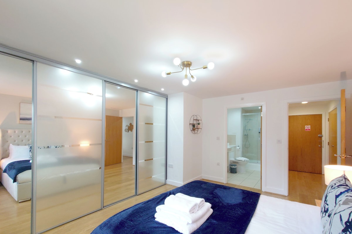 Habitación de hotel con cama, separador de vidrio, ducha y puerta que conduce a otra habitación.