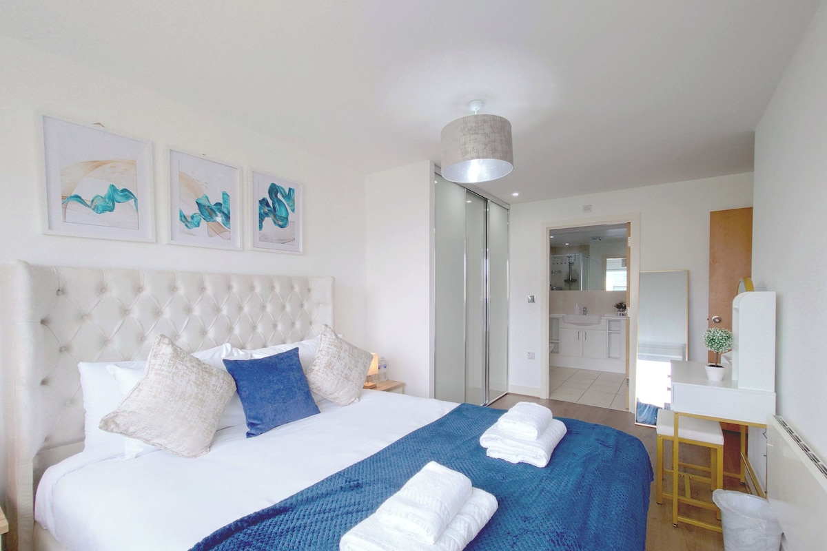Un dormitorio con cabecera capitonada, cama con ropa de cama blanca, almohadas azules y una manta azul. Hay tres obras de arte enmarcadas en la pared, una mesita de noche con una lámpara, y un baño visible a través de una puerta abierta.