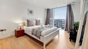 A modern bedroom with a bed, bedside table, and a large window with curtains. - Capital Stay - Two Bed Apartment In Excel (London)