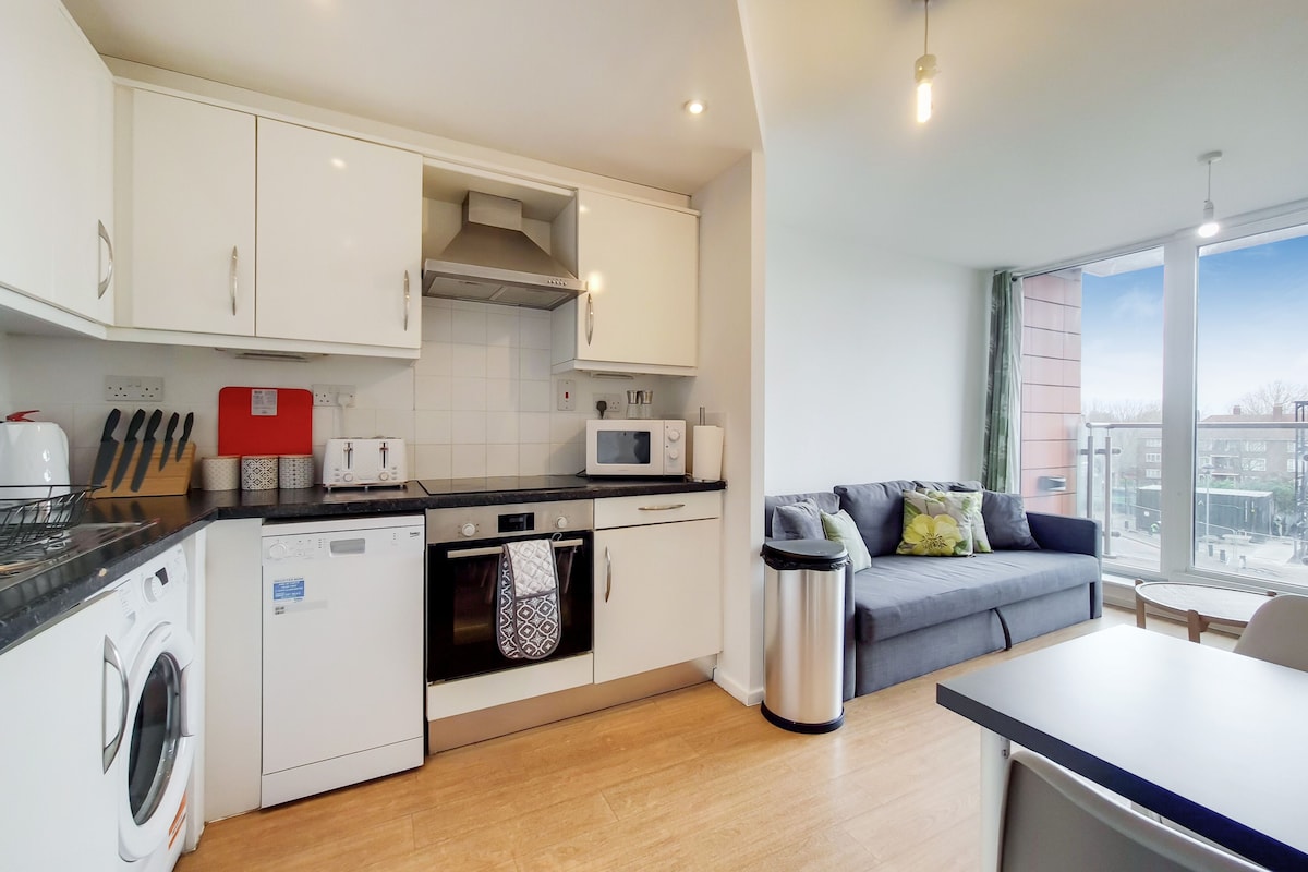 A compact kitchen with a microwave, oven, and washing machine, a grey sofa, and a large window.