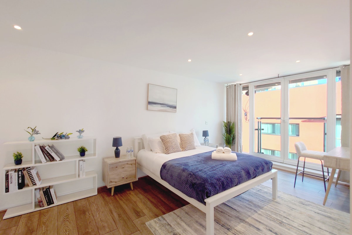 A modern hotel room with a bed, bedside tables, a bookshelf, a window with a view, and a balcony.