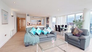 A modern living room with a sofa, armchairs, a glass coffee table, and a dining area with chairs and a table. - Capital Stay - Two Bed Apartment In Excel (London)