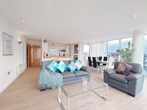 A modern living room with a sofa, armchairs, a glass coffee table, and a dining area with chairs and a table.