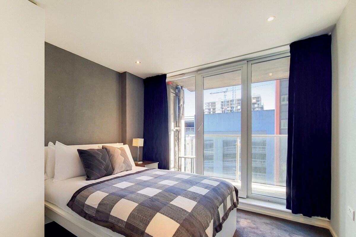 A bedroom with a bed, a nightstand, a lamp, a window with curtains, and a view of buildings.