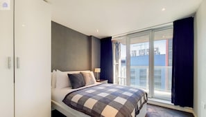 A bedroom with a bed, a nightstand, a lamp, a window with curtains, and a view of buildings.