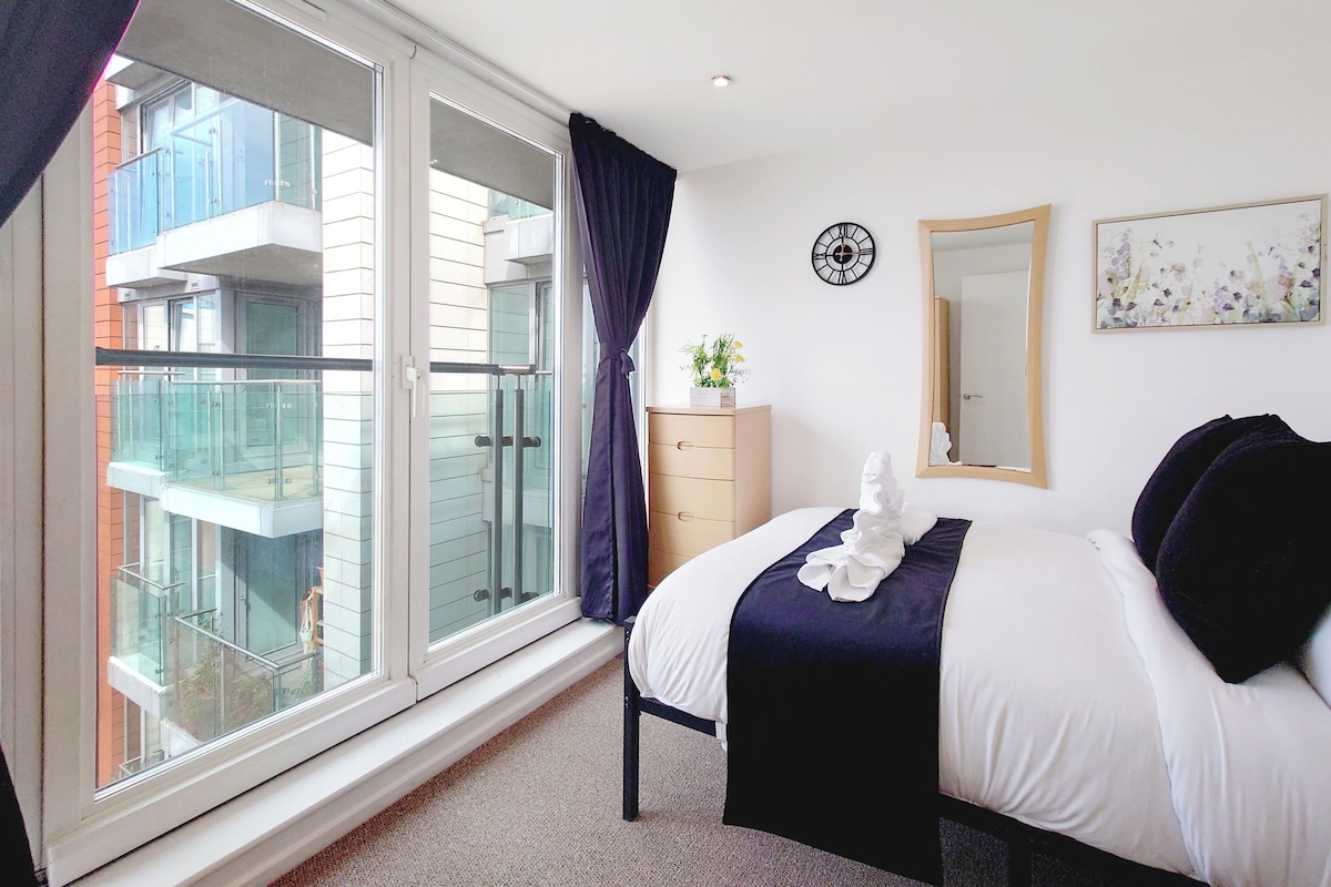 A hotel room with a large window, a bed with white and dark bedding, a dresser, a mirror, and a clock on the wall.