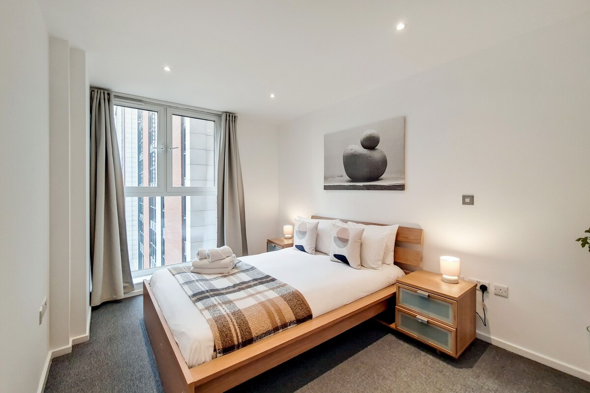 A modern hotel room with a large bed, bedside tables, a window with curtains, and a wall-mounted artwork.