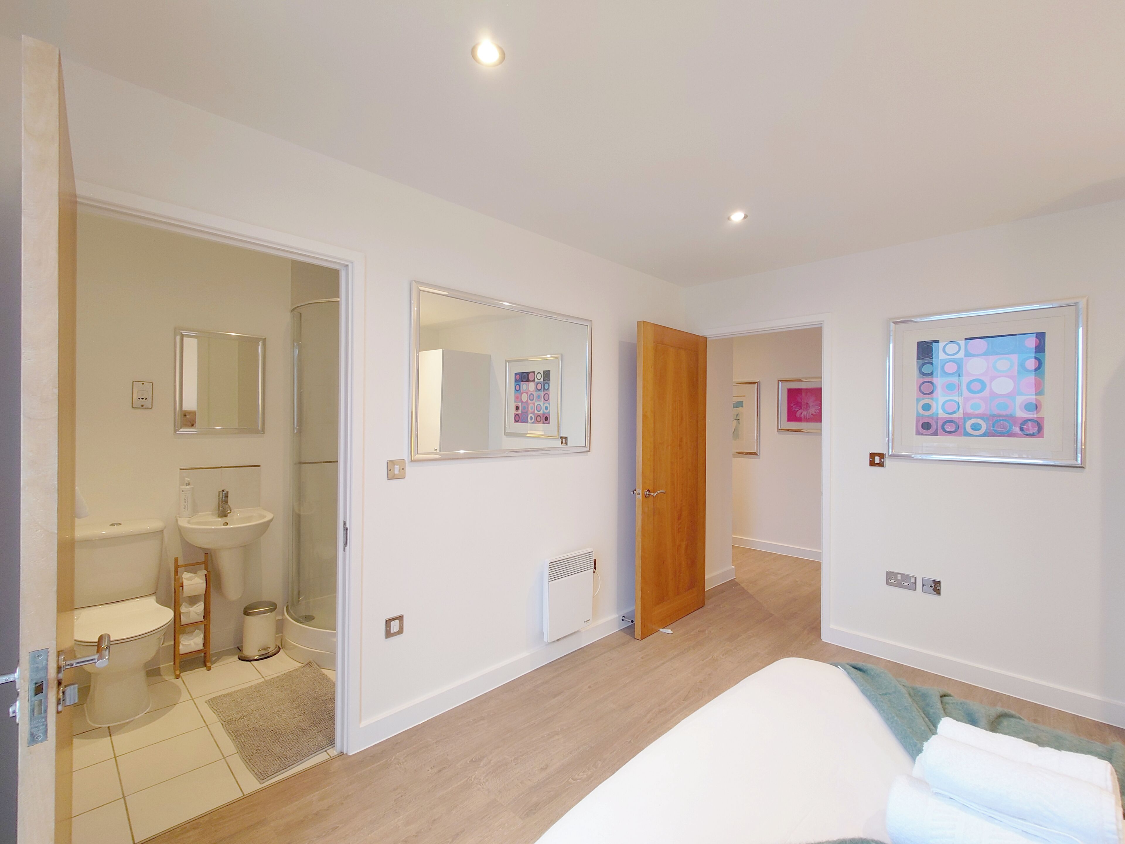 A modern bedroom with a bed, a bathroom with a toilet, sink, and mirror, and a hallway leading to another room.