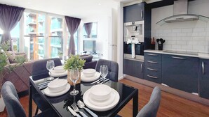 A modern dining area with a table set for six, a kitchen with dark cabinets and white countertops, and large windows with curtains. - Capital Stay - Two Bed Apartment In Excel (London)