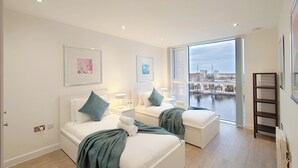 A modern bedroom with two beds, a large window with a city view, and a balcony. - Capital Stay - Two Bed Apartment In Excel (London)