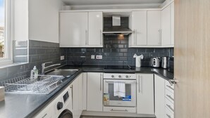 A modern kitchen with white cabinets, a stainless steel range hood, a built-in oven, and a dishwasher. - Two Bed Serviced Apartment (London)