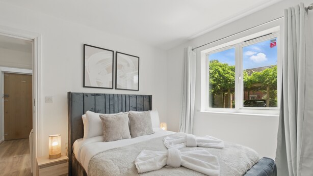 A bedroom with a bed, bedside lamps, a window with curtains, and framed artwork on the wall. - Two Bed Serviced Apartment (London)