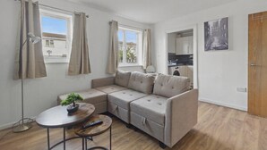 A modern living room with a sectional sofa, a small round table with a remote control, a floor lamp, and a view of the kitchen through an open door. - Two Bed Serviced Apartment (London)