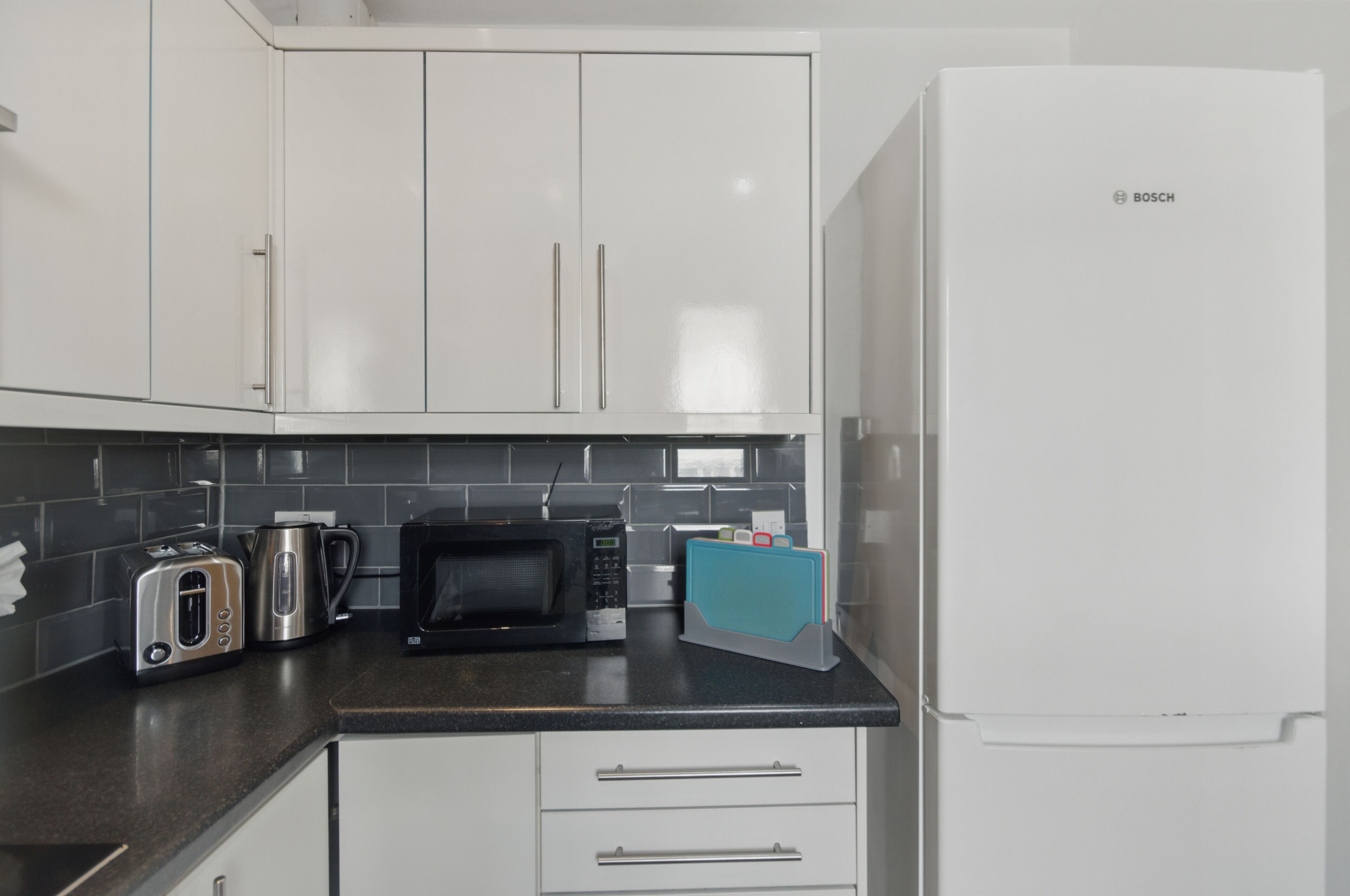 A kitchen with white cabinets, a Bosch refrigerator, a toaster, a kettle, and a microwave.