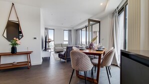 A modern dining area with a wooden table, cushioned chairs, a side table with a vase, and a large mirror on the wall. - Capital Stay - Three Bed Apartment In Excel (London)