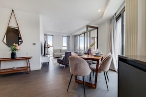 A modern dining area with a wooden table, cushioned chairs, a side table with a vase, and a large mirror on the wall. - Capital Stay - Three Bed Apartment In Excel (London)
