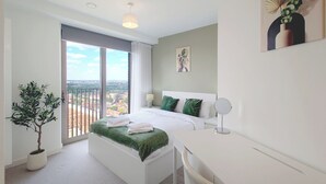 A modern hotel room with a bed, desk, and a large window offering a city view. - Capital Stay - Three Bed Apartment In Excel (London)