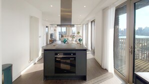 A modern kitchen with a central island, a dining area with a table and chairs, and a view of the city through large windows. - Capital Stay - Three Bed Apartment In Excel (London)