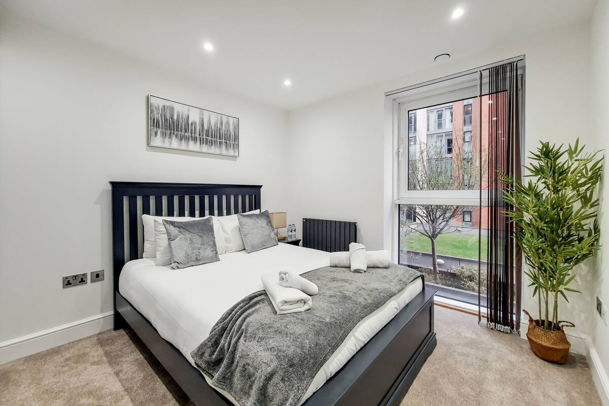 A modern bedroom with a large bed, a view of buildings outside, a potted plant, and a painting on the wall.