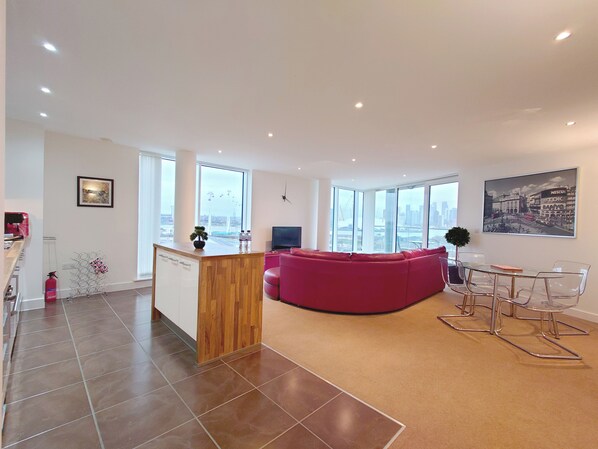 A modern living room with a wooden island, a red sofa, a glass dining table, and large windows offering a city view. - Capital Stay - Three Bed Apartment In Excel (London)