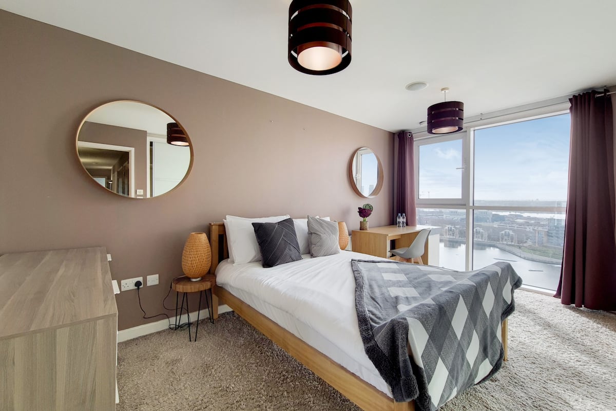 A modern hotel room with a large bed, two side tables, a round mirror, and a view of a cityscape through large windows.