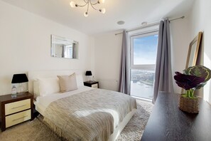 A hotel room with a bed, a nightstand, a lamp, a mirror, a window with curtains, and a view of a cityscape. - Capital Stay - Three Bed Apartment In Excel (London)
