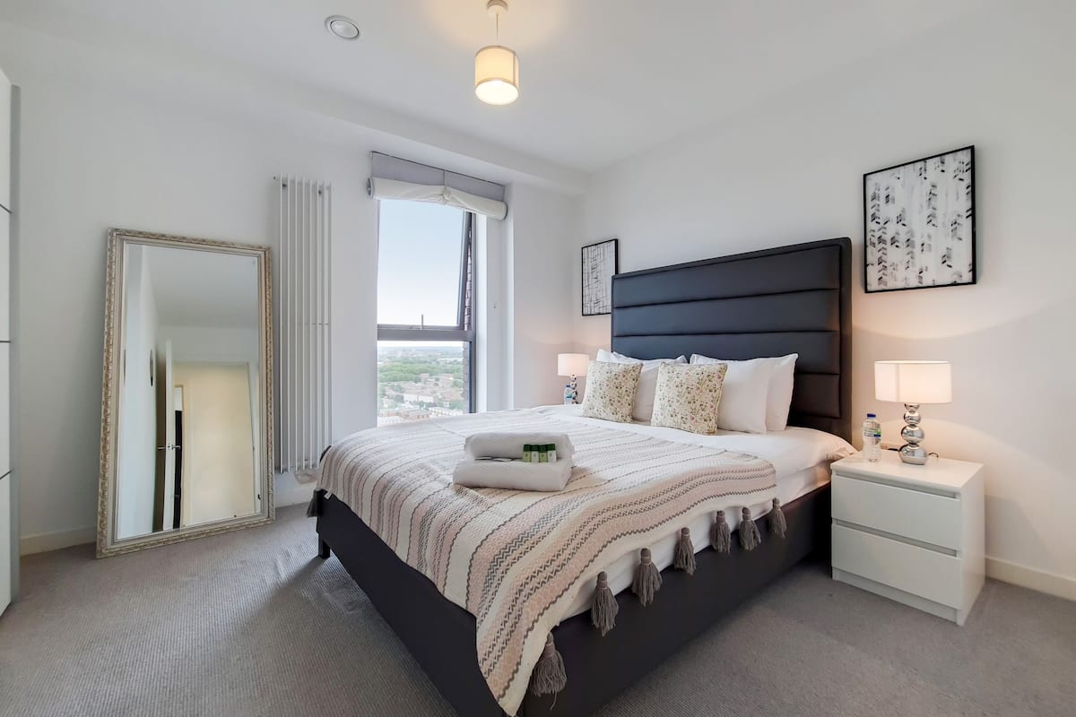 A hotel room with a large bed, a bedside table with a lamp, a mirror, and a window with a view.