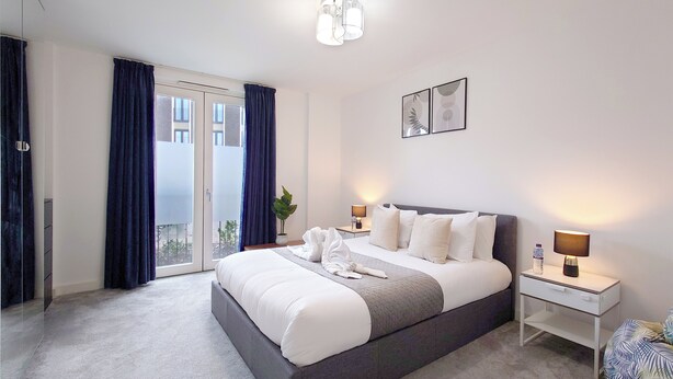 A modern hotel room with a large bed, a bedside table with a lamp, a potted plant, and a view through the glass door. - Luxury One Bed Apartment Near Olympic Park (London)