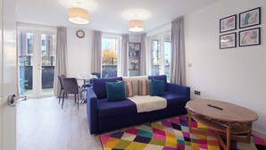 A living room with a sofa, a coffee table, a dining table with chairs, and a balcony with curtains. - Luxury One Bed Apartment Near Olympic Park (London)