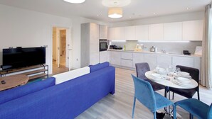 A modern living room with a flat-screen TV, a blue sofa, and a dining area with a table set for four. - Luxury One Bed Apartment Near Olympic Park (London)