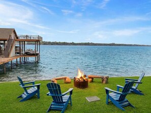A lakeside deck with a fire pit, blue Adirondack chairs, and a dock extending into the water. - Cedar Creek Lake Escape • Lakefront + Private Dock (Mabank)