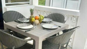 Un coin repas avec une table dressée pour deux personnes, entouré de chaises en osier, d’un panier de fruits et d’une fenêtre avec des stores.