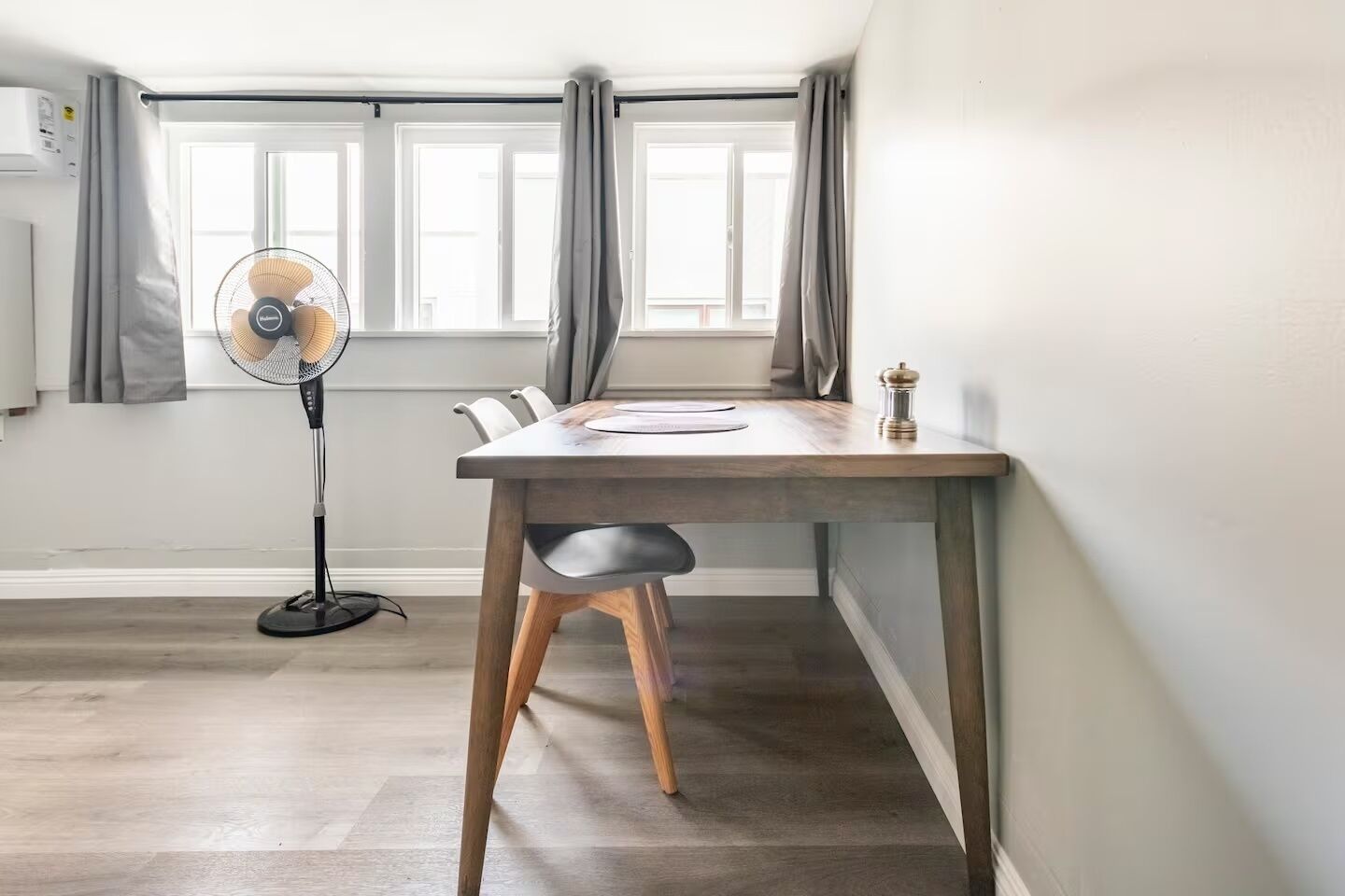 Un comedor con una mesa, sillas, un ventilador de pie y una ventana con cortinas.
