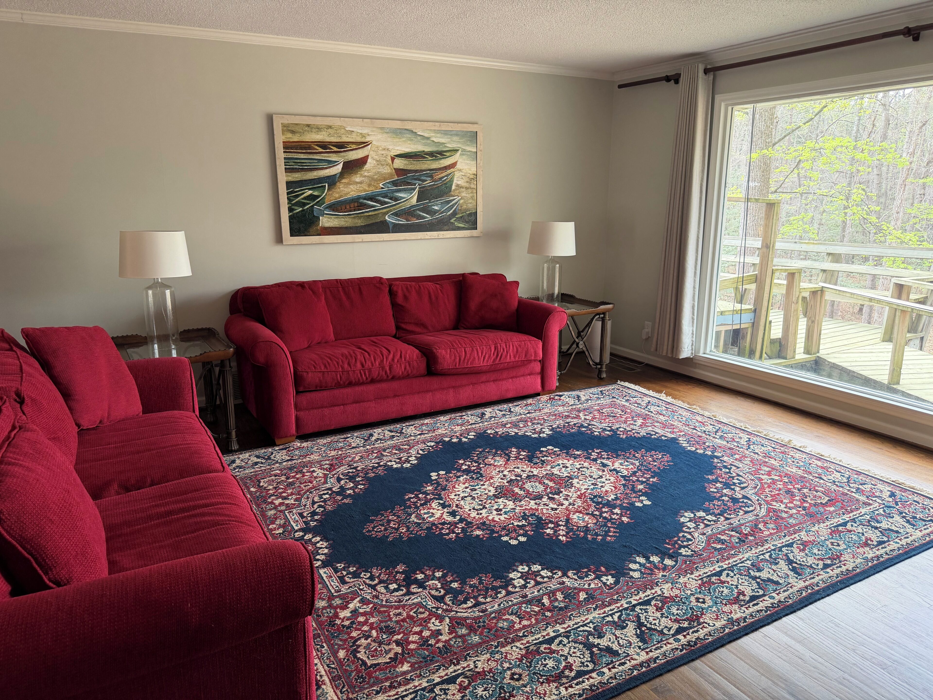 A living room with a red sofa, a patterned rug, a painting, and a view through a large window.