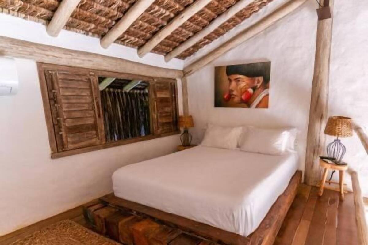 Un dormitorio con estructura de cama de madera, colchón blanco, cabecero de madera, una mesita de noche con lámpara, una ventana con contraventanas de madera y un cuadro en la pared.