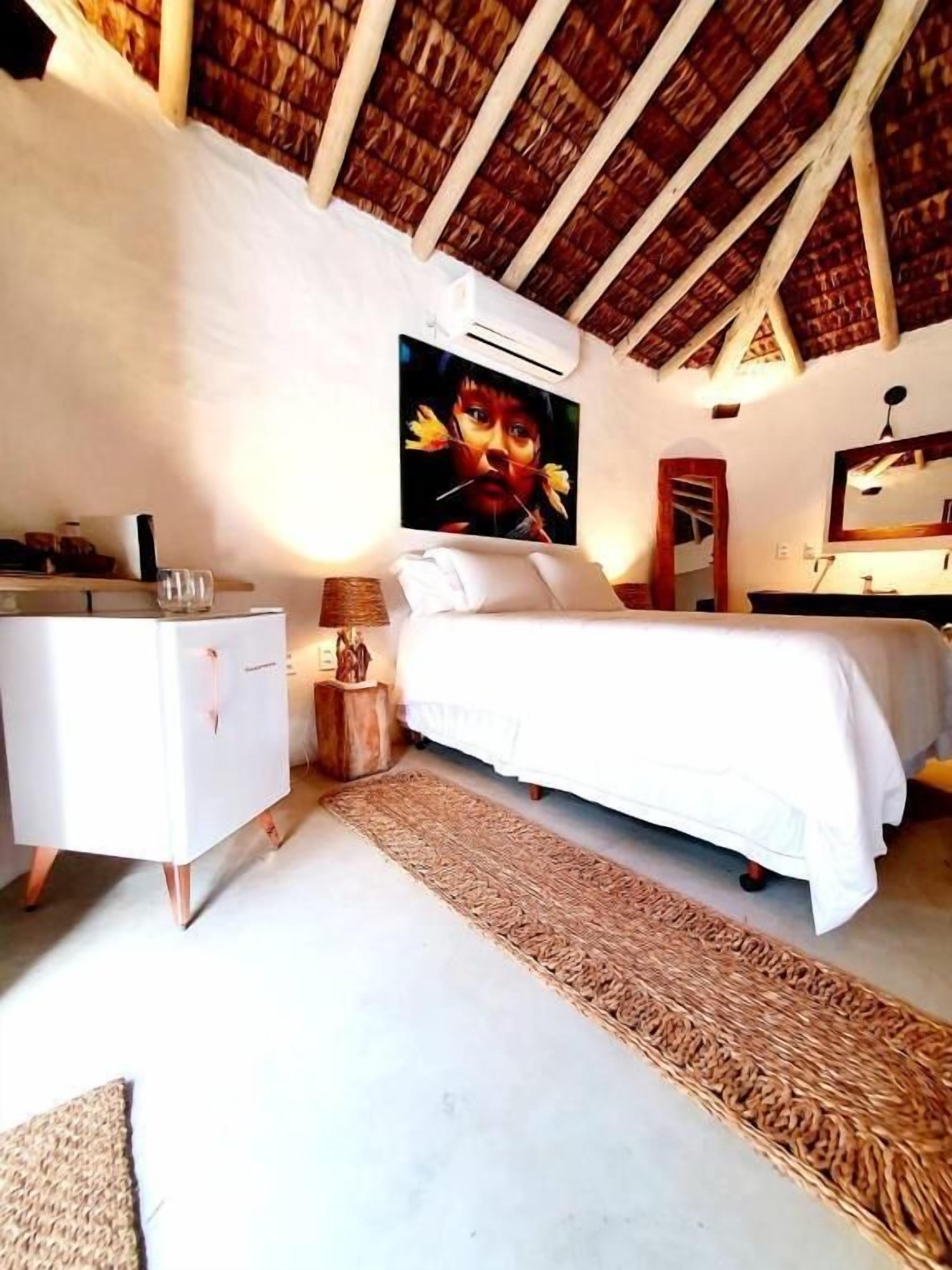 A bedroom with a bed, a nightstand, a dresser, a woven rug, a painting, and a ceiling with wooden beams.