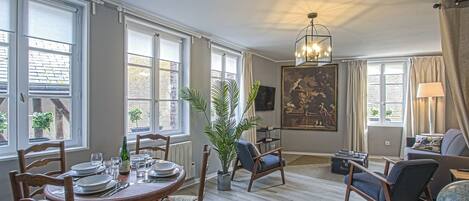 Ett rymligt vardagsrum med en matplats, stora fönster, en kristallkrona och en tavla på väggen.