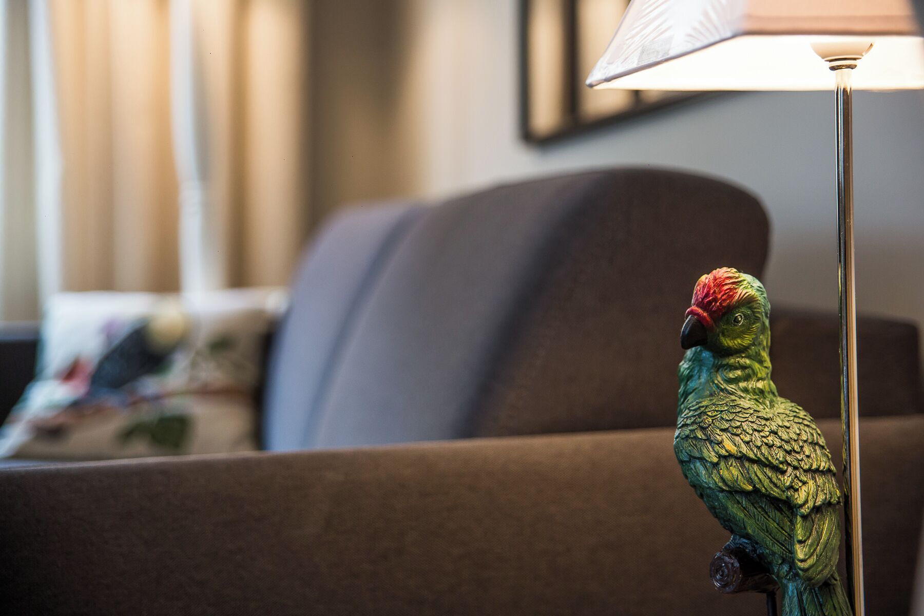 A parrot on a brown sofa with a lamp in the foreground.