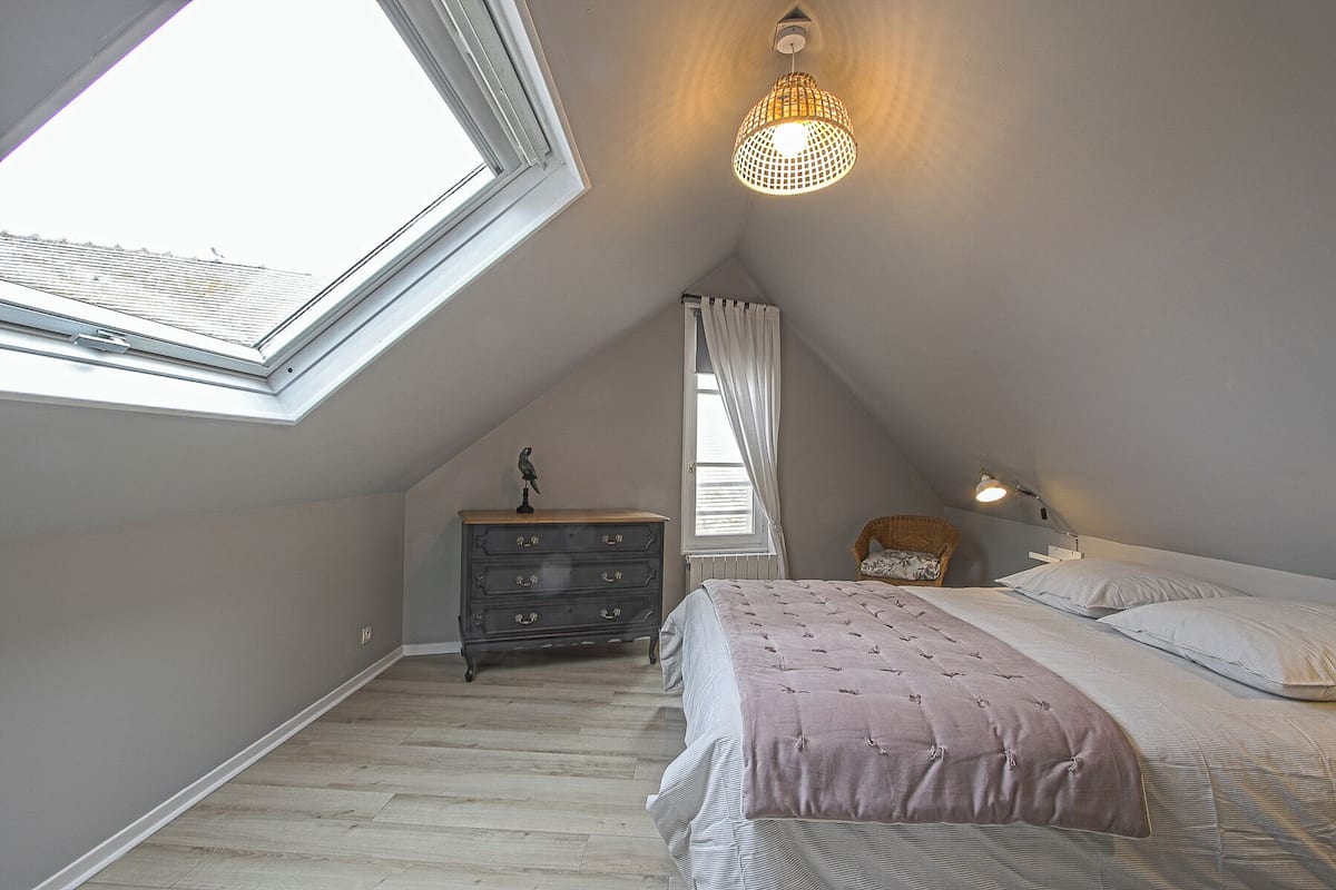 Une chambre avec un puits de lumière, une commode, un lit recouvert d’une couverture rose et un luminaire suspendu.