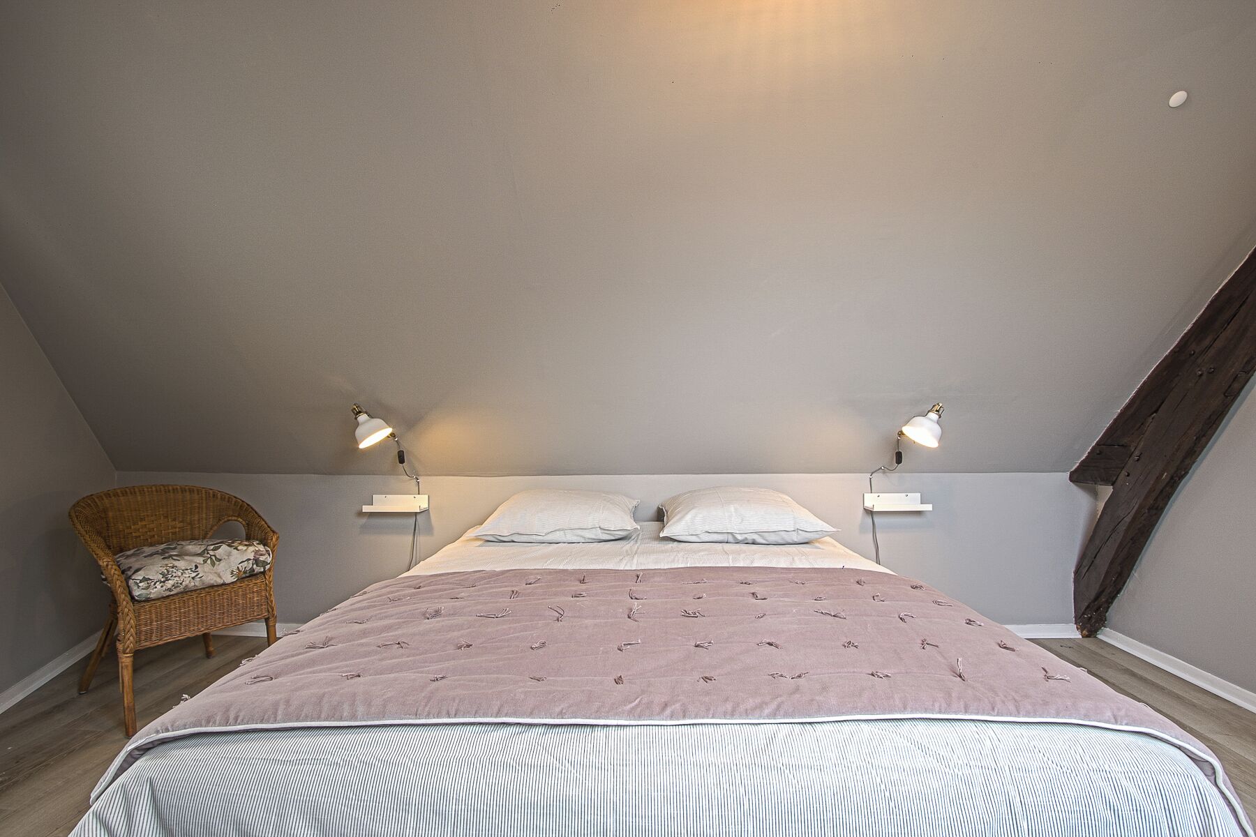 A bedroom with a bed, two bedside lamps, a wicker chair, and a sloped ceiling.