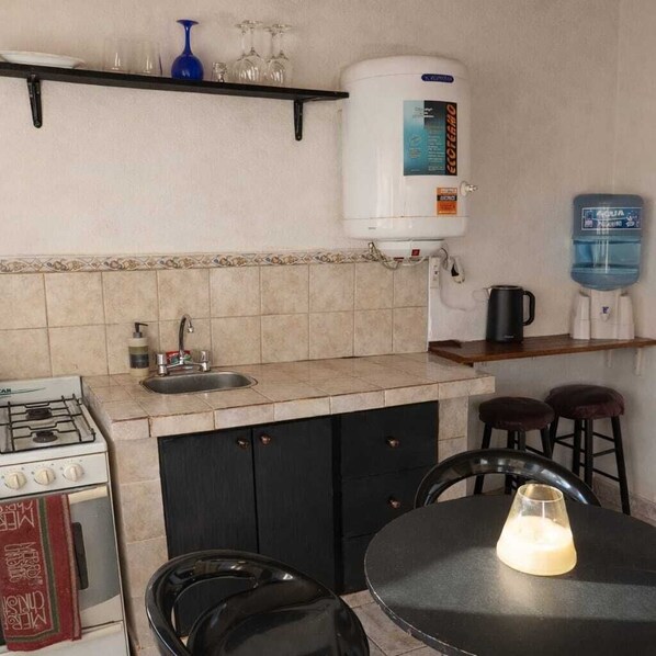 Una cocina con estufa a gas, pileta y dispensador de agua. Hay dos taburetes y una mesa con una vela.