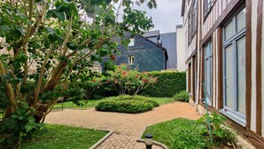 A courtyard with a brick pathway, green hedges, and a tree with pink flowers. - La Venelle Saint-Nicolas (Rouen)