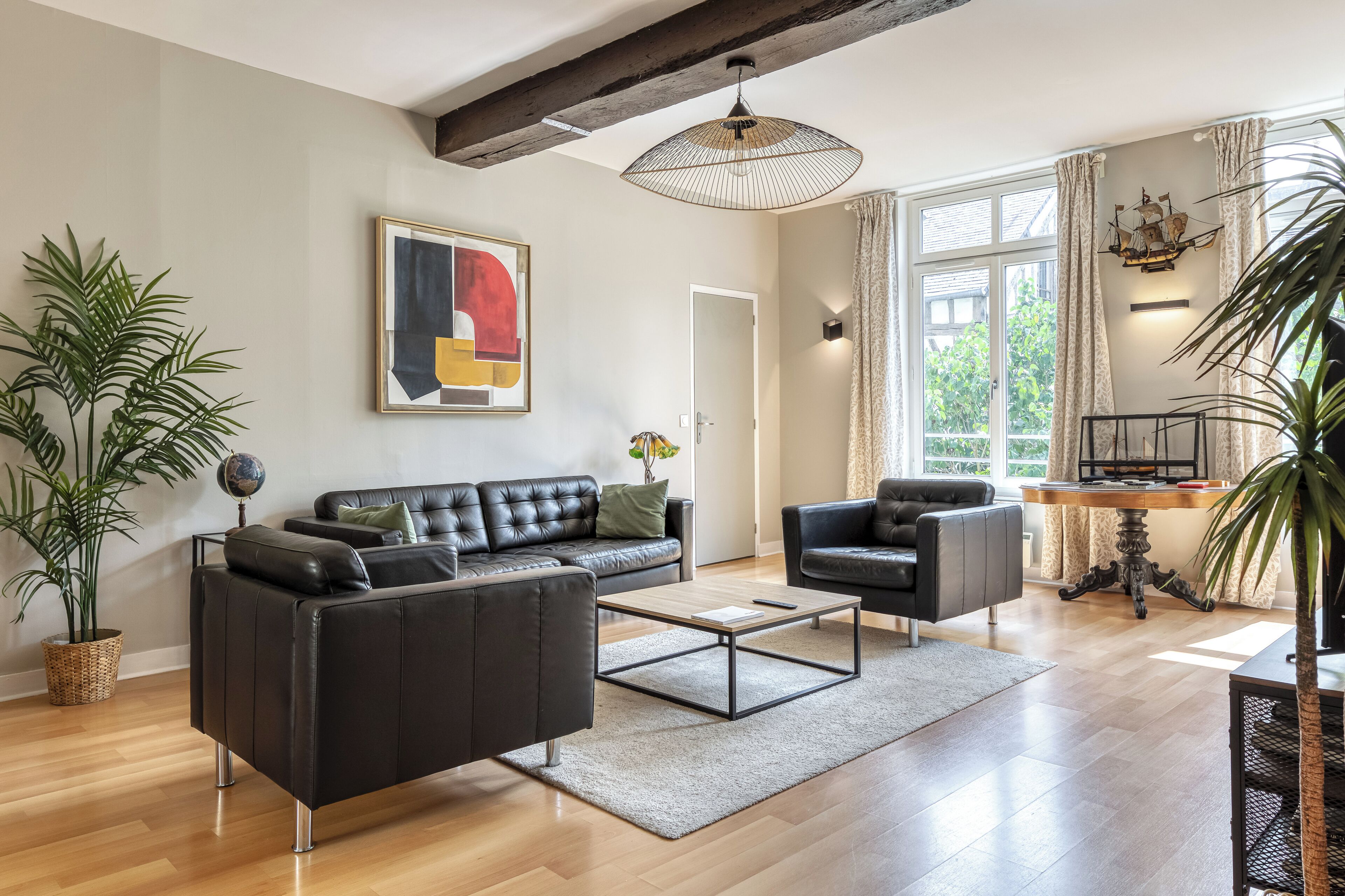 Un salon moderne comprenant un canapé, une table basse et des fauteuils, une table à manger décorée avec un motif de bateau, et une grande fenêtre avec des rideaux.