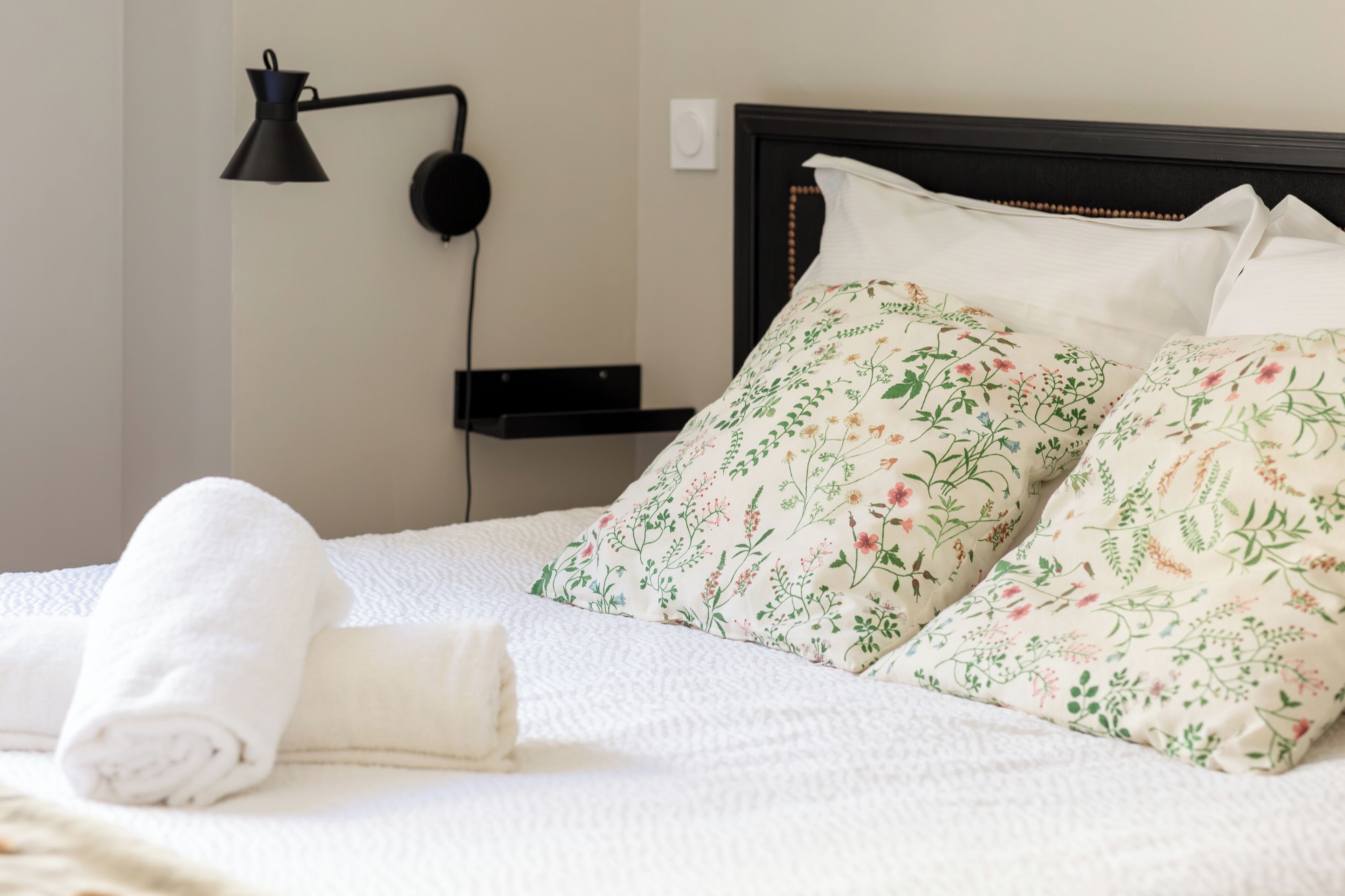 A neatly made bed with floral pillows, a rolled white towel, a black desk lamp, and a wall-mounted speaker.