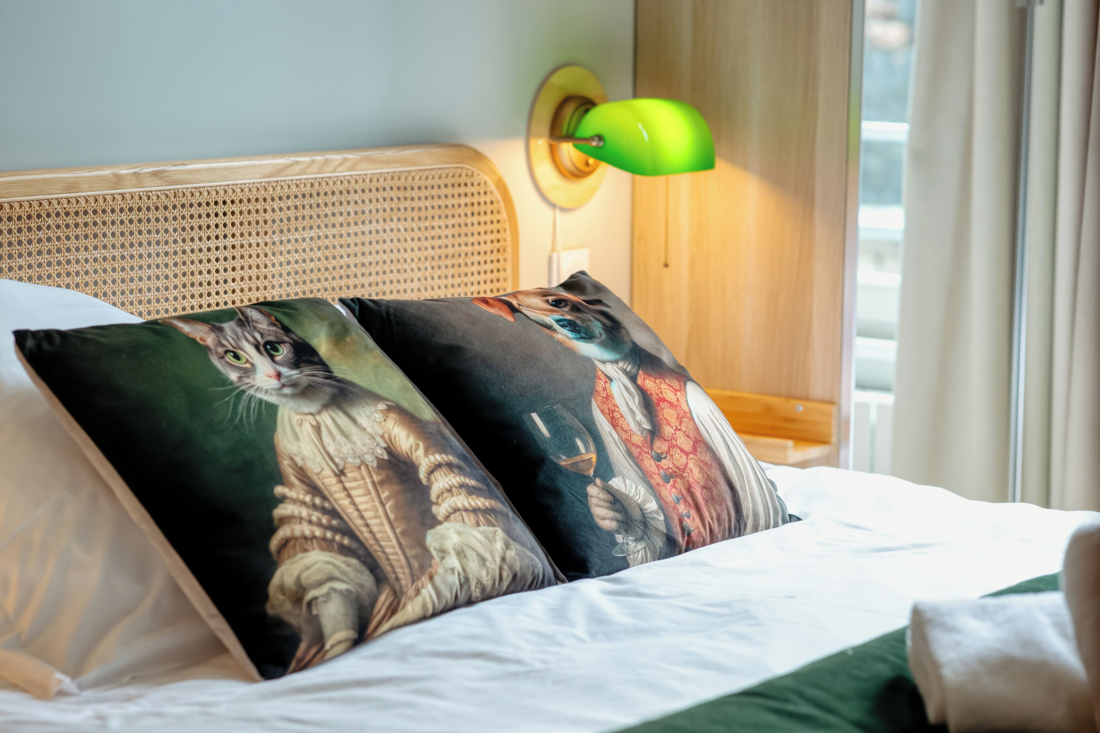 Una cama con dos almohadas decoradas con ilustraciones de gatos, una lámpara de pared verde y un cabecero de madera.