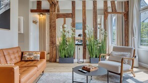 A living room with a sofa, armchair, and coffee table, wooden beams, potted plants, and a painting on the wall. - La Venelle Saint-Nicolas (Rouen)