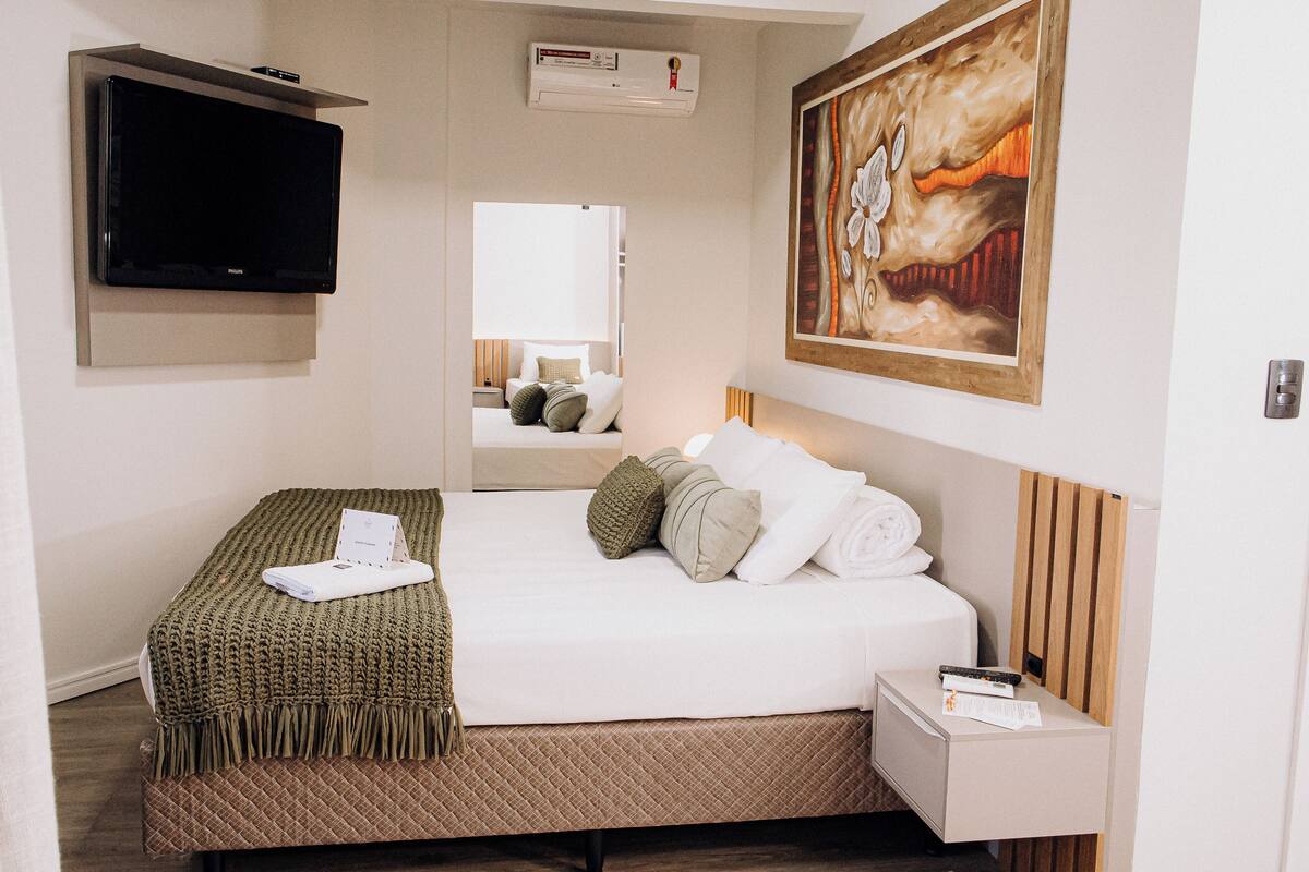 Habitación de hotel con cama, mesita de noche, televisor montado en la pared, un cuadro y un equipo de aire acondicionado.