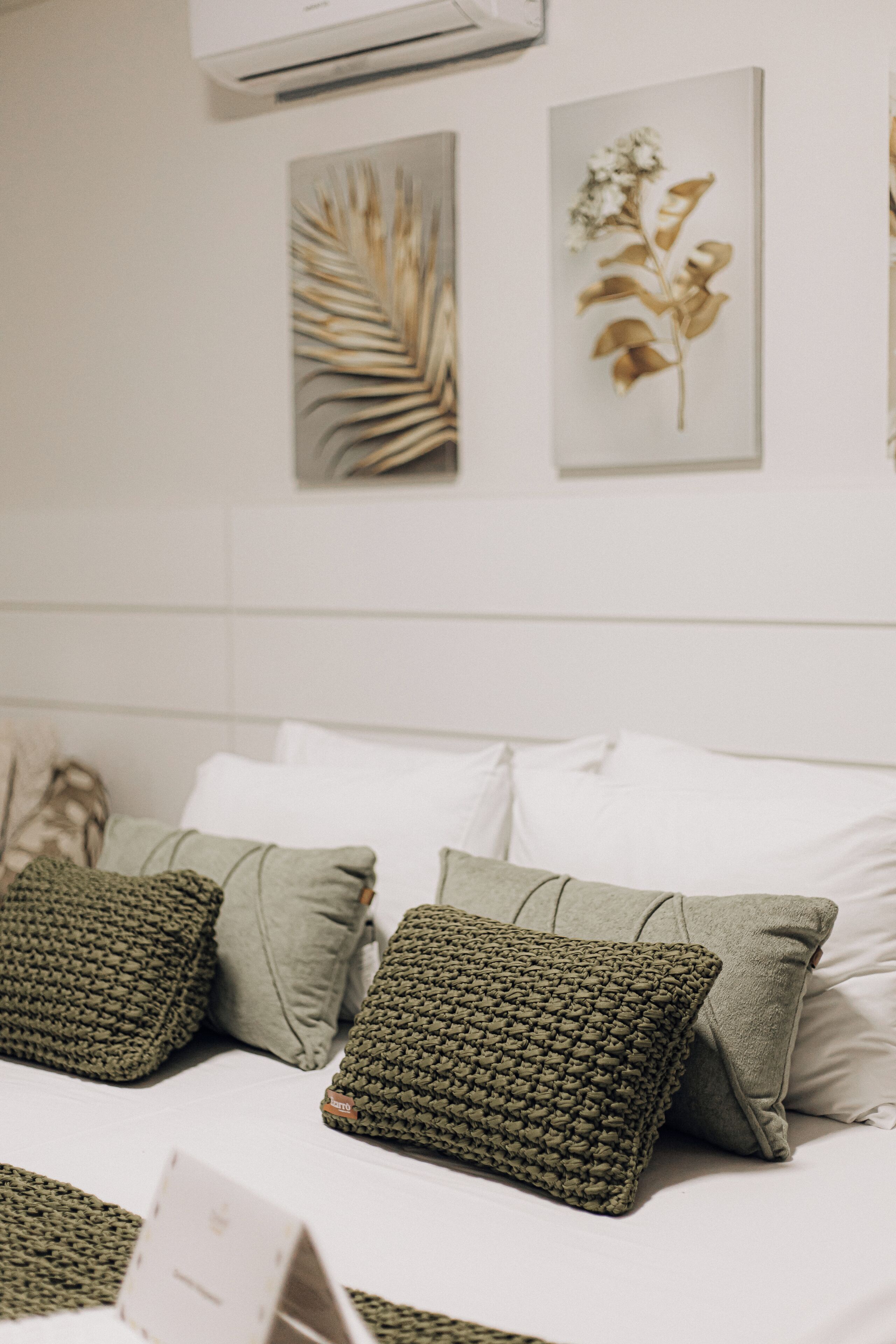 A bed with white linens and green pillows, a wall-mounted air conditioner, and two framed botanical prints.